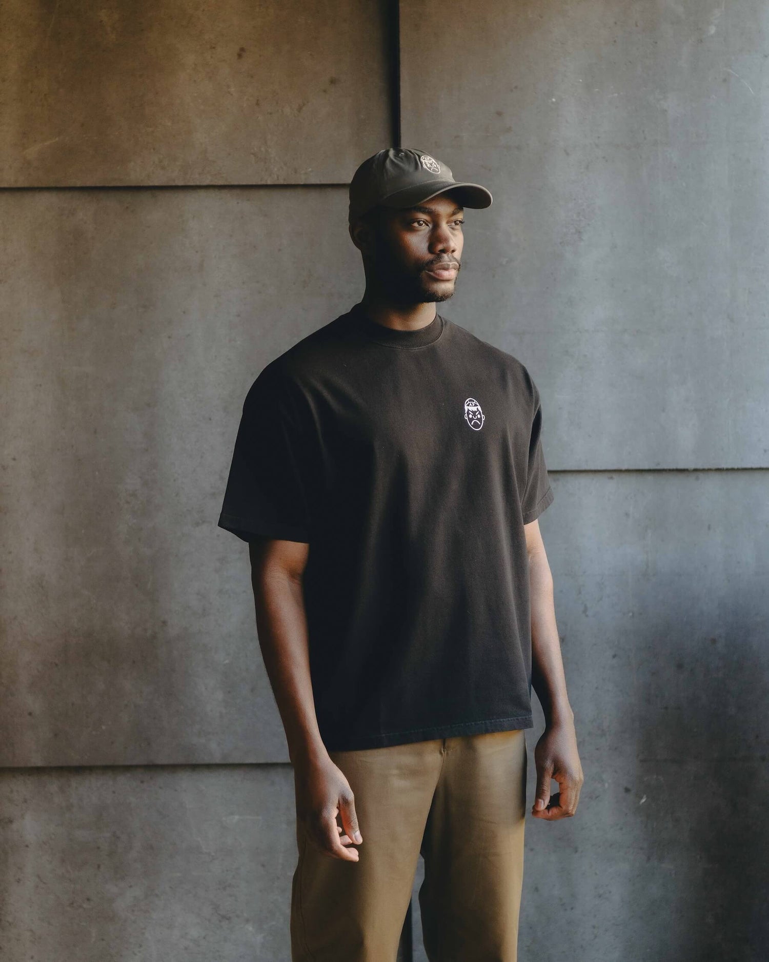 A man wearing a black heavy oversized t-shirt and an Angry Pablo performance cap, in front of a grey slate background.